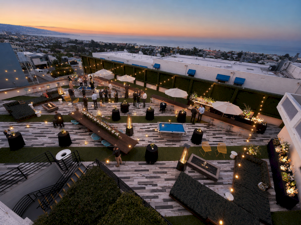 A lit rooftop terrace hosts an evening event with tables and guests, overlooking the ocean at sunset.