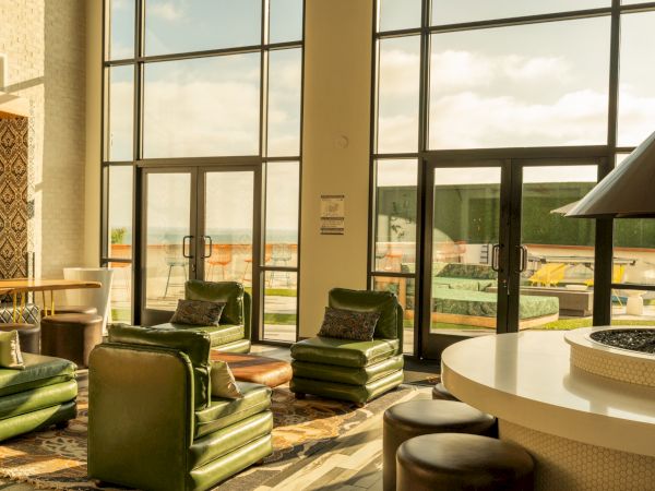 A cozy lounge with green chairs, large windows, and a view of outdoor furniture on a sunny day. A round fireplace adds warmth.
