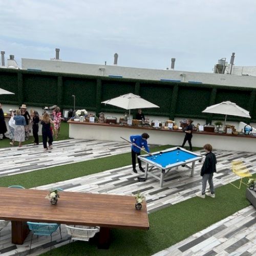 An outdoor area with people, a pool table, tables, and chairs, under umbrellas on a wooden deck with a green backdrop.