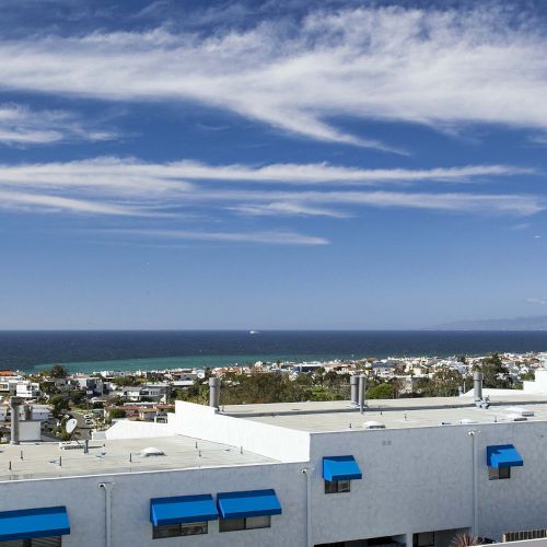 The image shows a bright day with a coastal cityscape, featuring rooftops, the ocean, distant mountains, and clouds above.