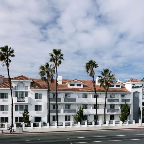 A white hotel with red-tiled roof, palm trees, and a view of the ocean under a partly cloudy sky by a road intersection.