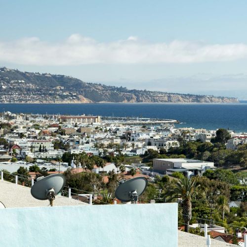 A coastal cityscape with rooftops, satellite dishes, and greenery, overlooking a bay and distant hills under a clear blue sky.