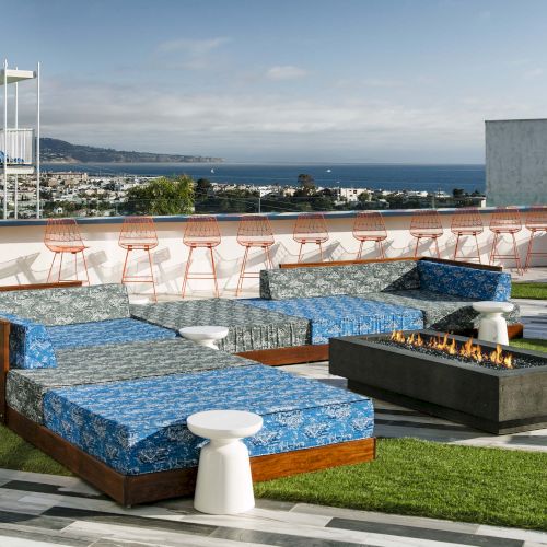 A rooftop patio with blue seating, fire pit, and stools overlooks a scenic ocean view with rows of pink chairs along the railing.