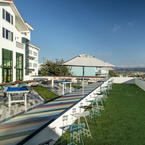A rooftop with tables, stools, and a sunshade by a building, overlooking a landscape under a clear sky.