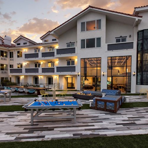 A modern hotel exterior at sunset with balconies, outdoor seating, and game tables in the foreground.
