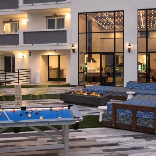 The image shows an outdoor area with a lit pool table, string lights, lounge seating, and fire pits, adjacent to a building with large windows.