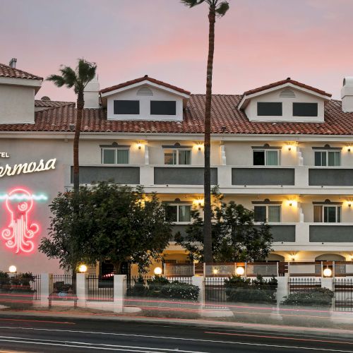 The image shows a hotel with "Hotel Hermosa" on a neon sign, surrounded by palm trees, at dusk.