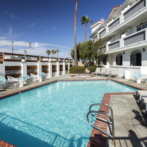 A hotel pool area with clear blue water, surrounded by lounge chairs and a white multi-story building. Sunny weather with a clear sky.
