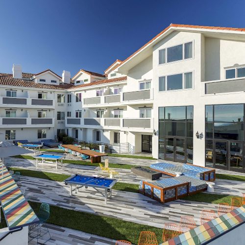 The image shows a modern white building with balconies, colorful seating, and tables in an outdoor area on a sunny day.
