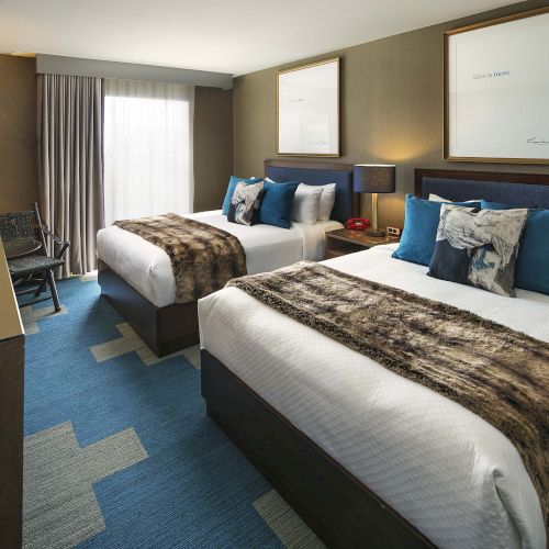 This image shows a hotel room with two double beds, a flat-screen TV, a large window, and modern decor featuring blue and brown tones.