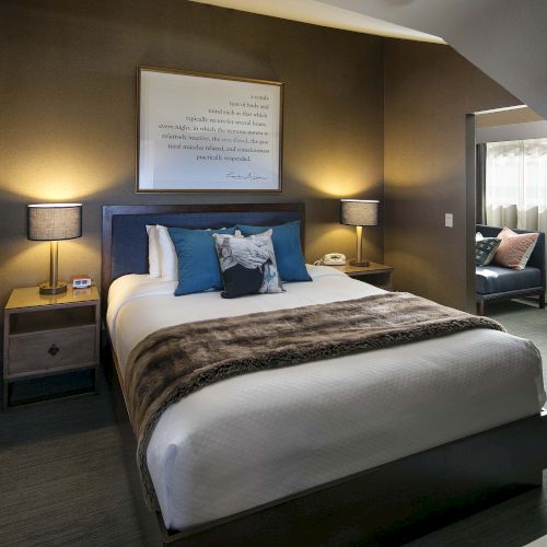 A cozy bedroom with a large bed, fur blanket, bedside lamps, art on the wall, and seating area by a window.