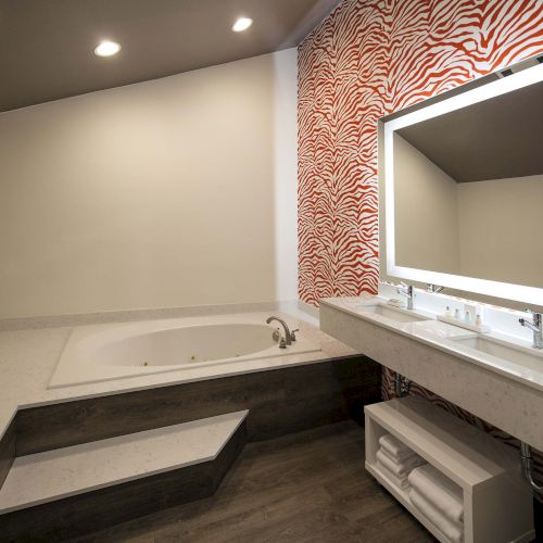 A modern bathroom with a corner bathtub, steps, a large mirror, vibrant red patterned wall, and towel storage beneath the sink area.