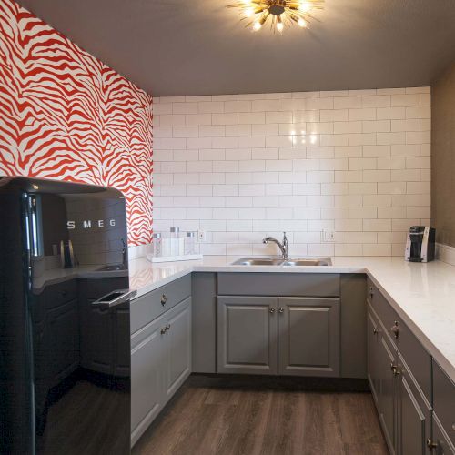 A kitchen with gray cabinets, white countertops, a black fridge, red zebra-patterned wall, and a modern light fixture on the ceiling.
