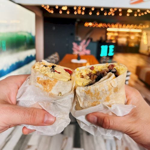 Two hands holding wrapped burritos or sandwiches inside a modern, brightly lit restaurant setting.