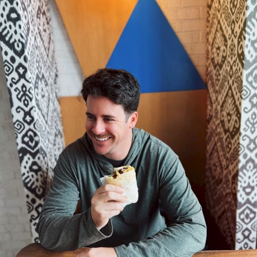 A person is sitting at a table, smiling while holding a sandwich or wrap, with patterned walls and a geometric design in the background.