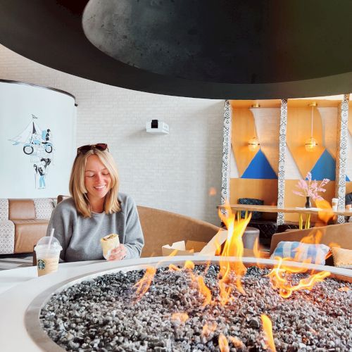 A person sits by a modern indoor fire pit, enjoying a drink. Cozy seating and decor create a warm atmosphere.