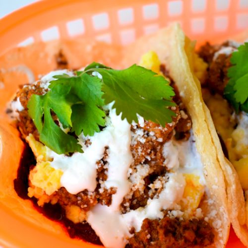 Two tacos with meat, cheese, cilantro, and sauce in a basket, garnished with fresh green herbs.