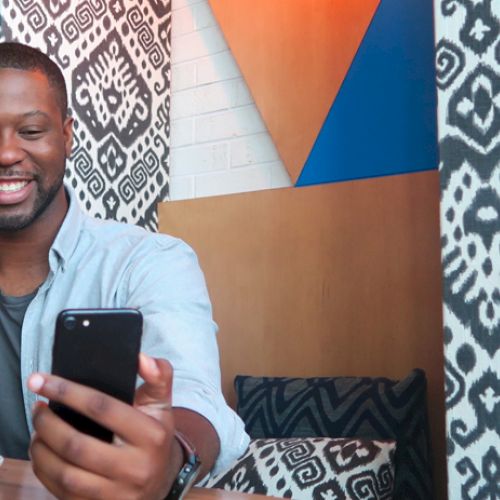A person is sitting at a table, smiling at a phone, holding a cup, with a muffin nearby, and patterned decor in the background.