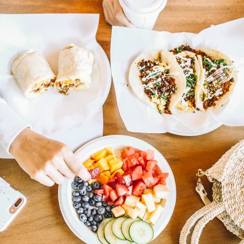 A table with tacos, a burrito, a plate of fruits and veggies, and people interacting. There's also a cup and a woven bag.