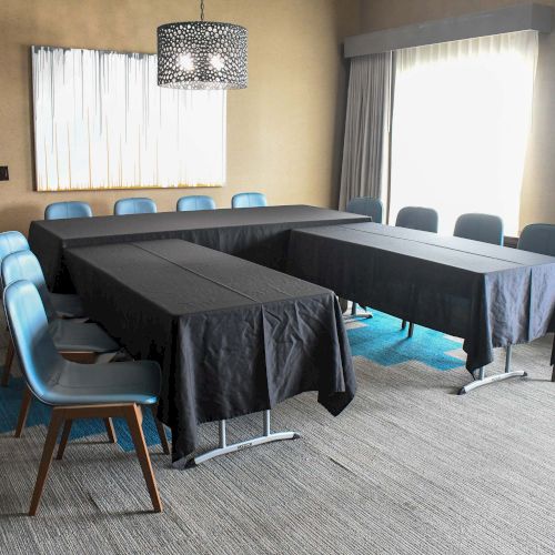 A conference room with U-shaped tables, black tablecloths, blue chairs, abstract wall art, and ample natural light streaming in.