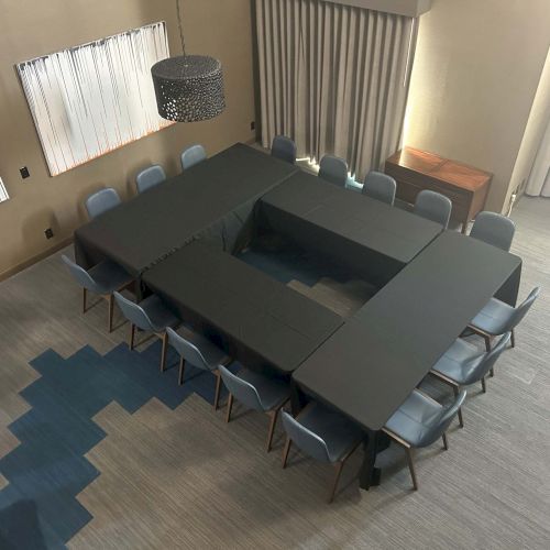 A conference room setup with tables arranged in a square U-shape, surrounded by chairs, and decorated with curtains and wall art.