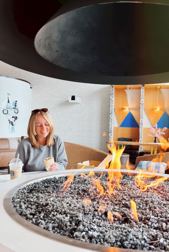 A person sitting at a table with drinks near a circular fire pit indoors, surrounded by modern decor and a cozy atmosphere.