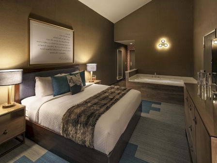 A cozy bedroom with a large bed, stylish lamps, and a wall-mounted TV. There's a framed quote and a visible bathtub area.