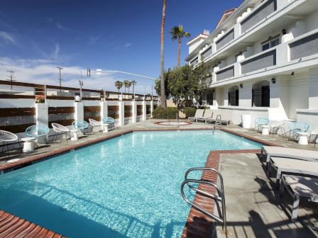 A hotel pool area with loungers, chairs, and a multi-story building in the background under a clear blue sky.