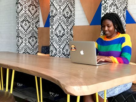 A person wearing a colorful sweater is using a laptop on a wooden table. The room has patterned decor and cushioned seating around.