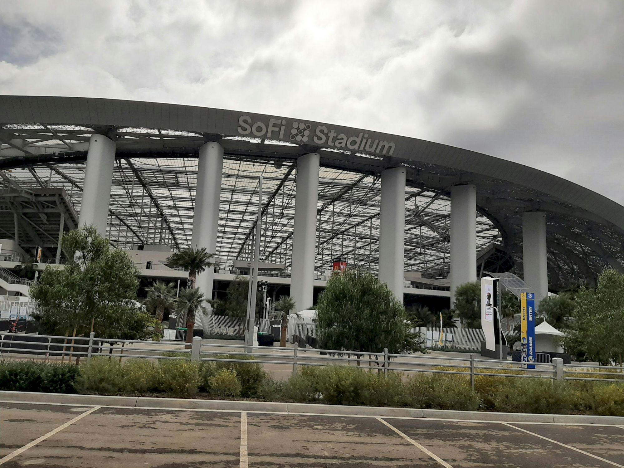 The image shows the exterior of SoFi Stadium, with its grand architectural design, surrounded by trees and an empty parking lot.