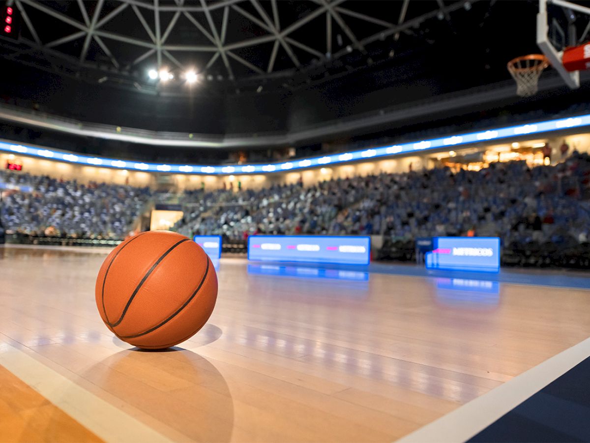 A basketball lies on the court of an empty arena, under bright lights, with spectators' seats in the background.