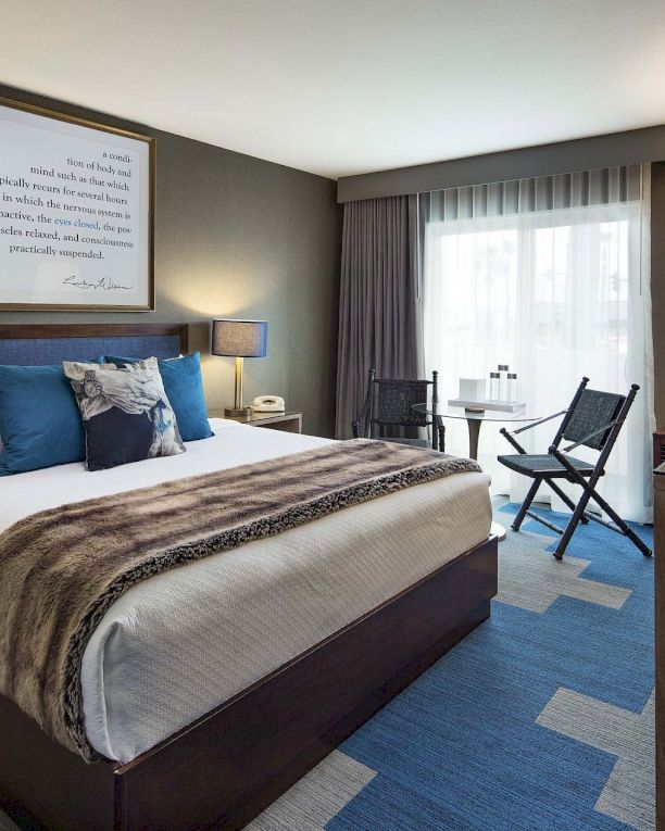 A modern hotel room with a king bed, two nightstands, blue accents, a desk, chair, and a bright window with sheer curtains. End.