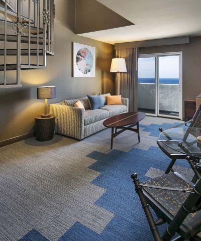 A cozy hotel suite with a living area, spiral staircase, blue zigzag carpet, sofa, coffee table, lamps, desk, and a balcony view.