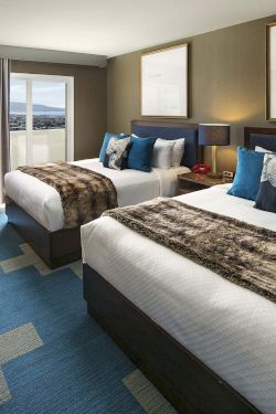 Two double beds in a modern hotel room with blue accents, a desk, wall art, a TV, and a window with curtains.