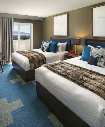 Two double beds in a modern hotel room with blue accents, a desk, wall art, a TV, and a window with curtains.