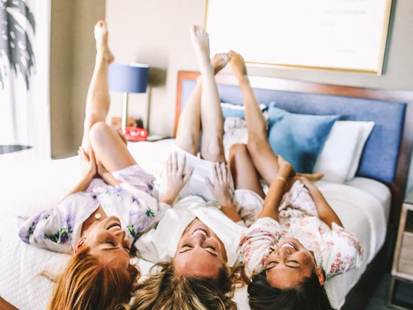Three friends lying on a bed, heads together, legs in the air, smiling and posing for a playful photo in a bright hotel room.