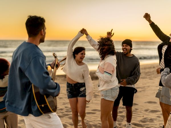 A group of friends on a beach at sunset, dancing and cheering, one playing a guitar as they celebrate together.