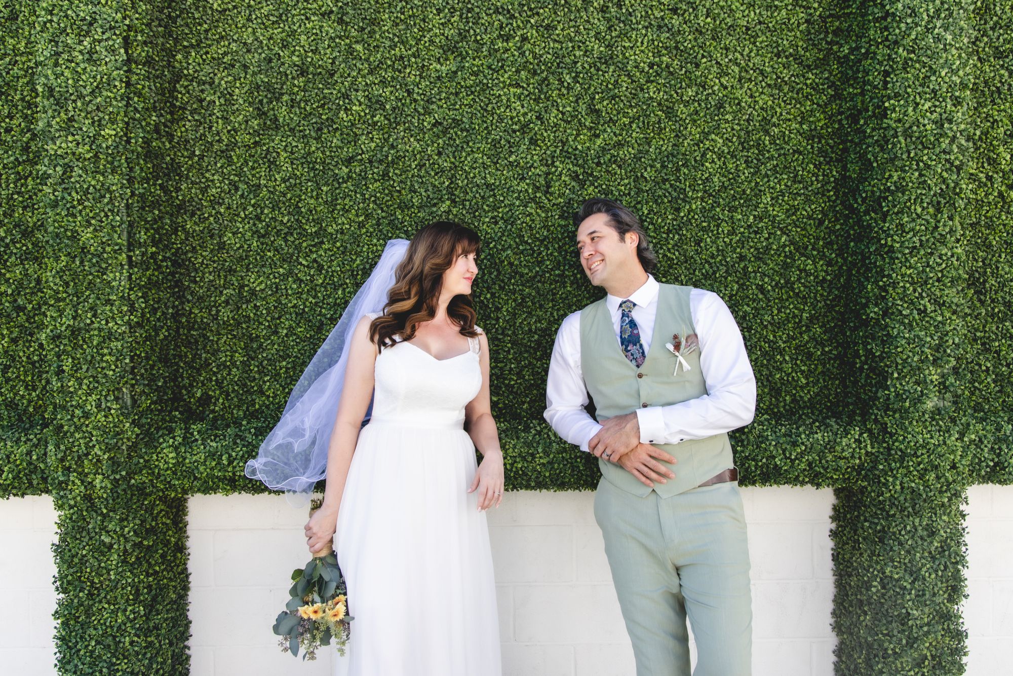 A bride and groom pose smiling in front of a lush green hedge wall, with the bride holding a bouquet.