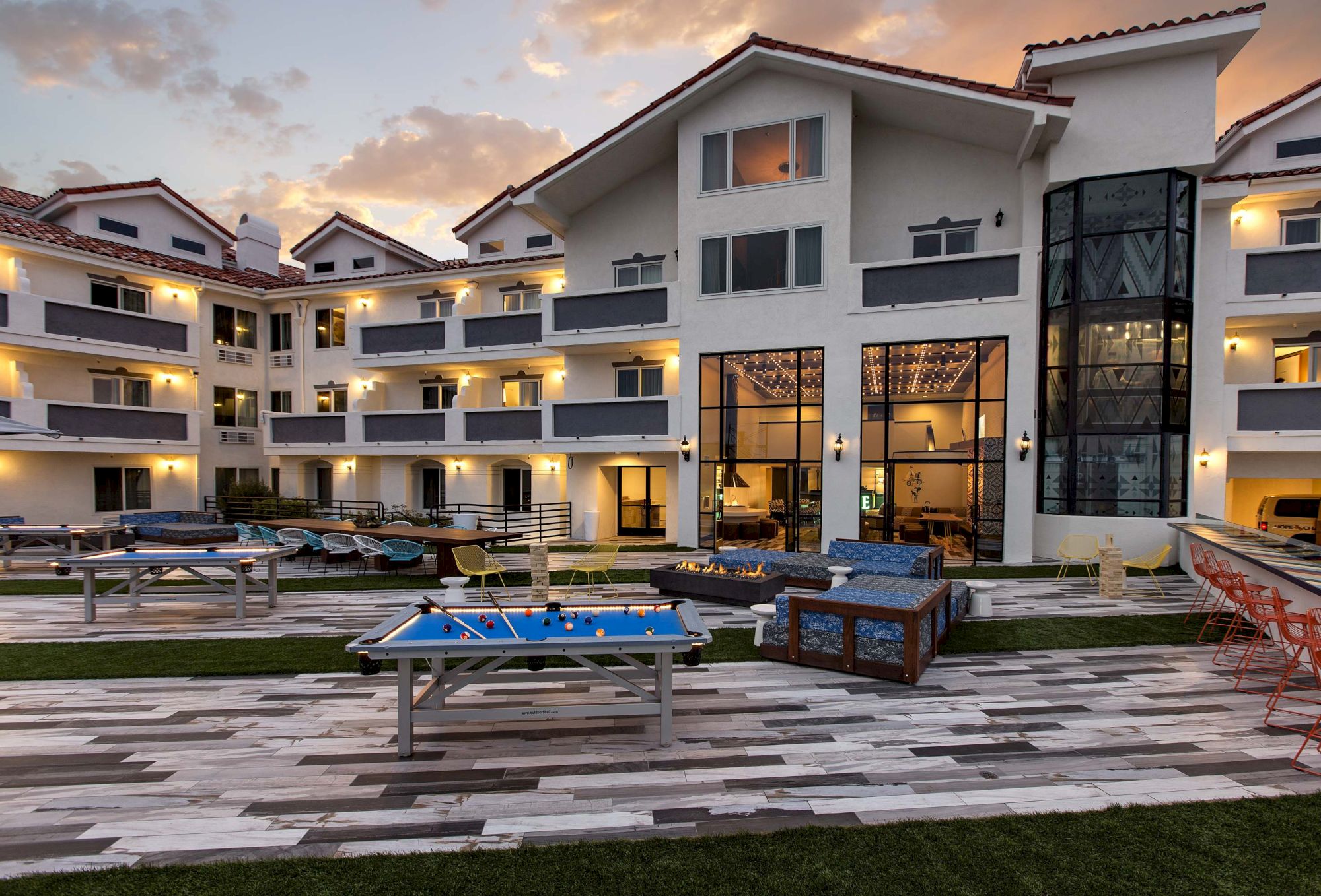 A modern apartment courtyard at dusk with a blue pool table, lounge seating, a fire pit, and lit patio umbrellas.
