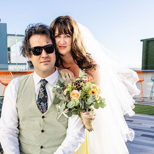 A bride and groom pose outdoors on a terrace, the bride holding a bouquet while the groom wears sunglasses and a suit vest.