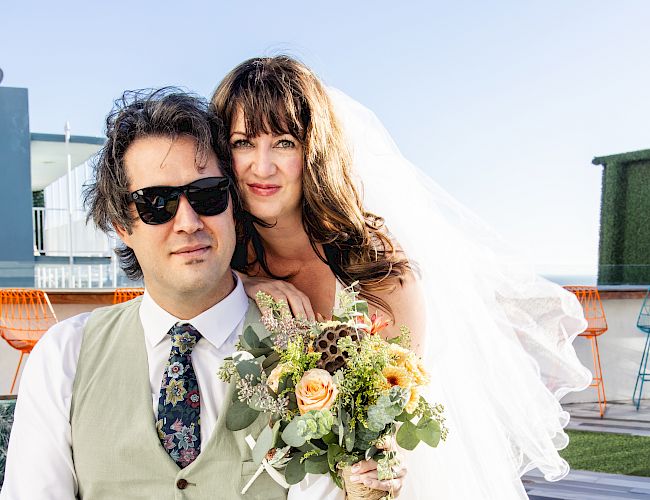 A bride and groom pose outdoors on a terrace, the bride holding a bouquet while the groom wears sunglasses and a suit vest.