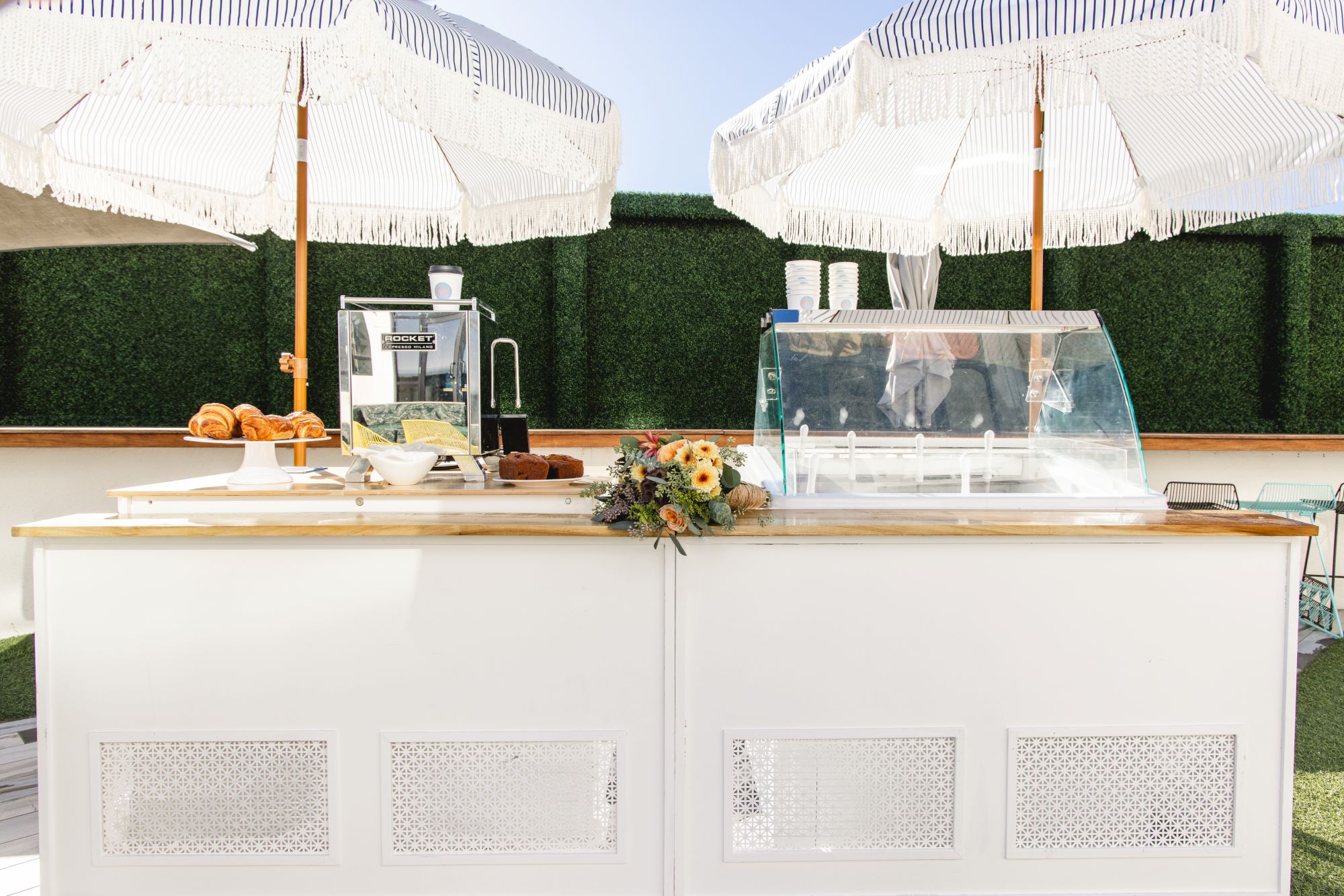 An outdoor catering buffet with croissants and bread, coffee cups, and a glass food display under white event tents.