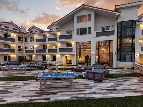 A modern apartment courtyard at dusk with patio lights, a pool table, lounge seating, and a fire pit in front of large windows.