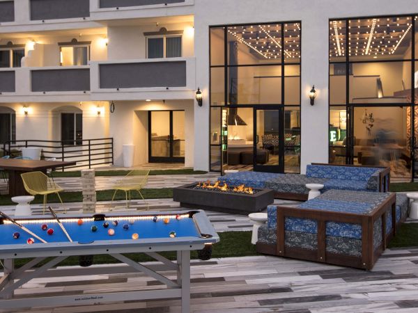 Outdoor patio at dusk with a billiards table, cushioned loungers, a lit fire pit, and string lights through glass doors.