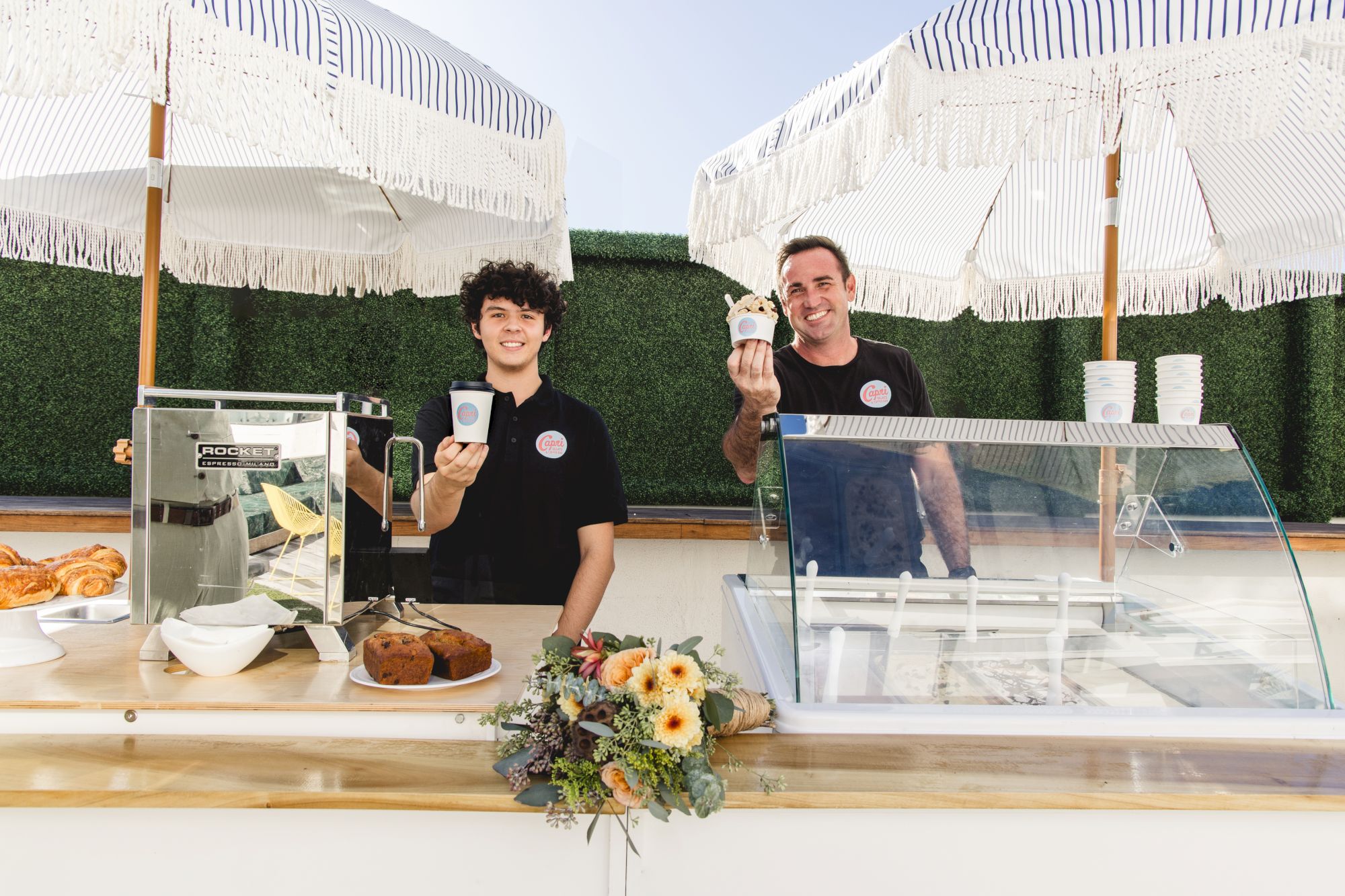 Two men in black shirts run an outdoor ice-cream cart under white striped tents, smiling and holding cone and cups.
