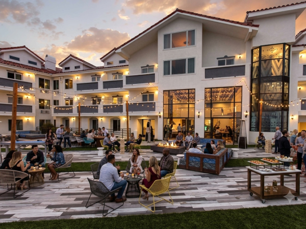 An evening outdoor courtyard party with string lights, guests chatting, and a buffet/food table in front of apartment buildings.